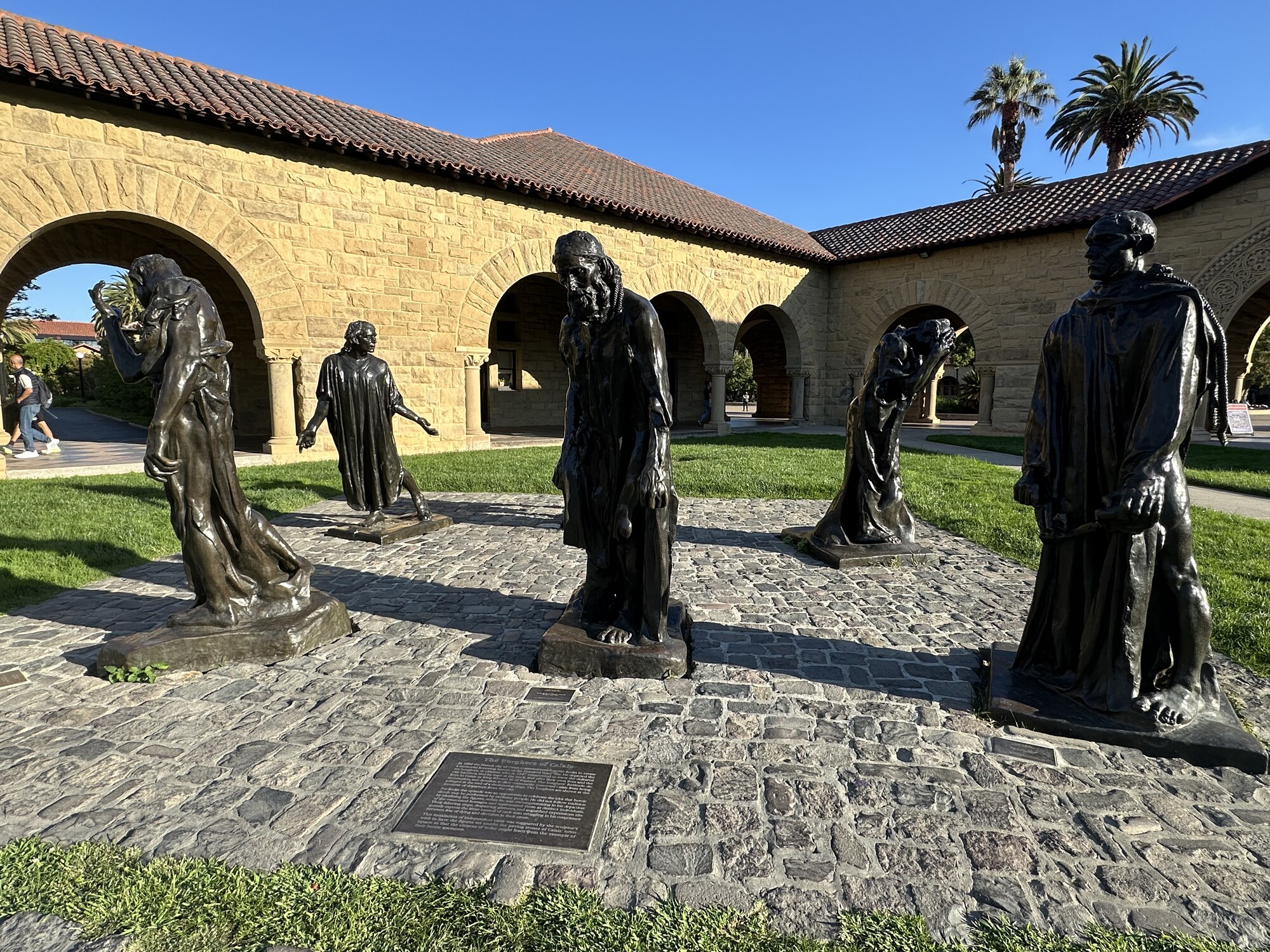Several bronze figures standing apart in a sunlit courtyard.
