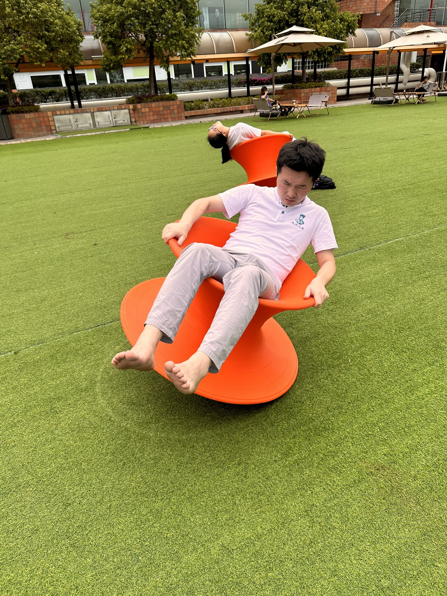 A barefoot person tilting on an orange chair on a green lawn.