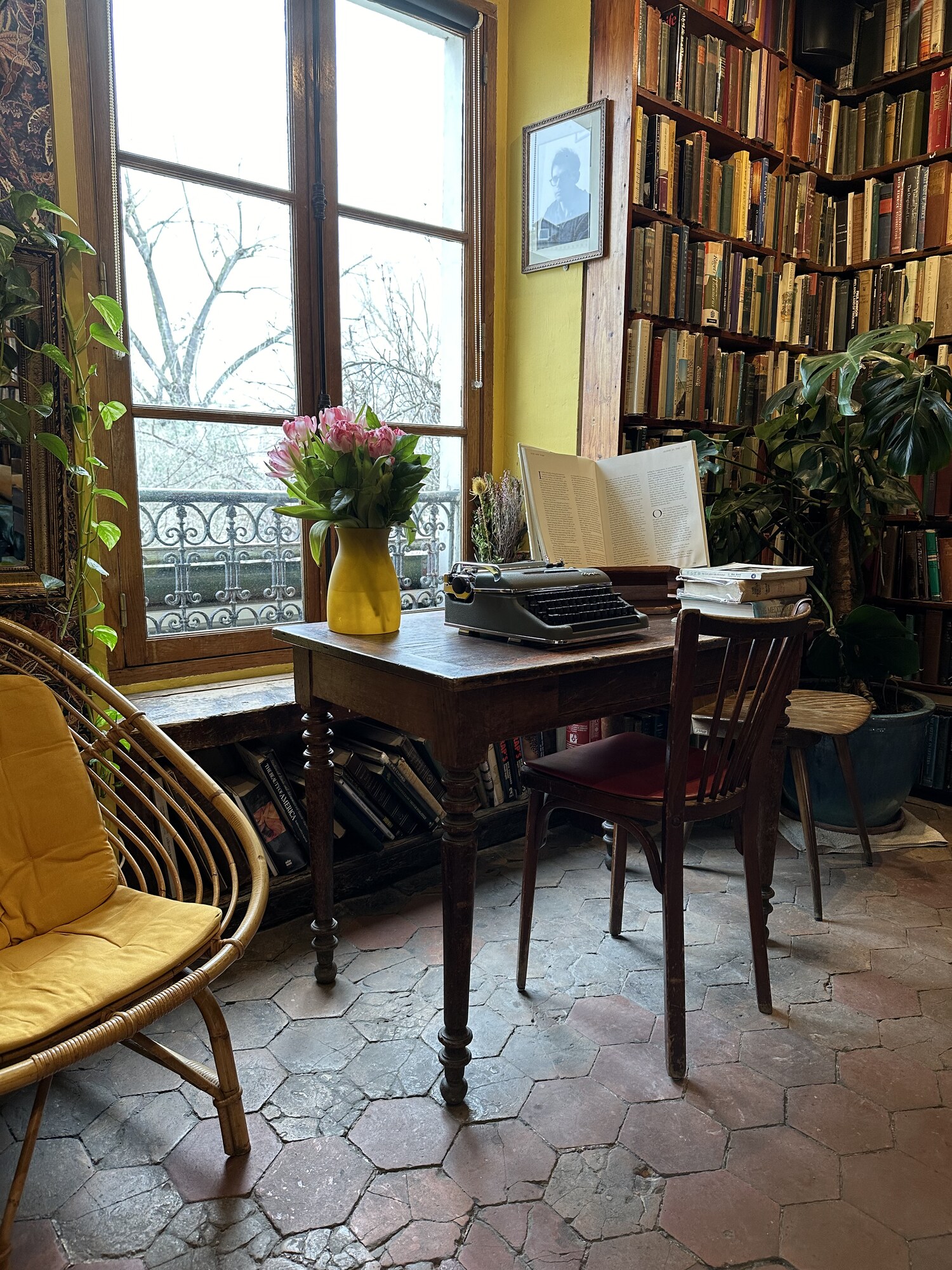 Books, a typewriter and flowers on a desk beside a tall window.