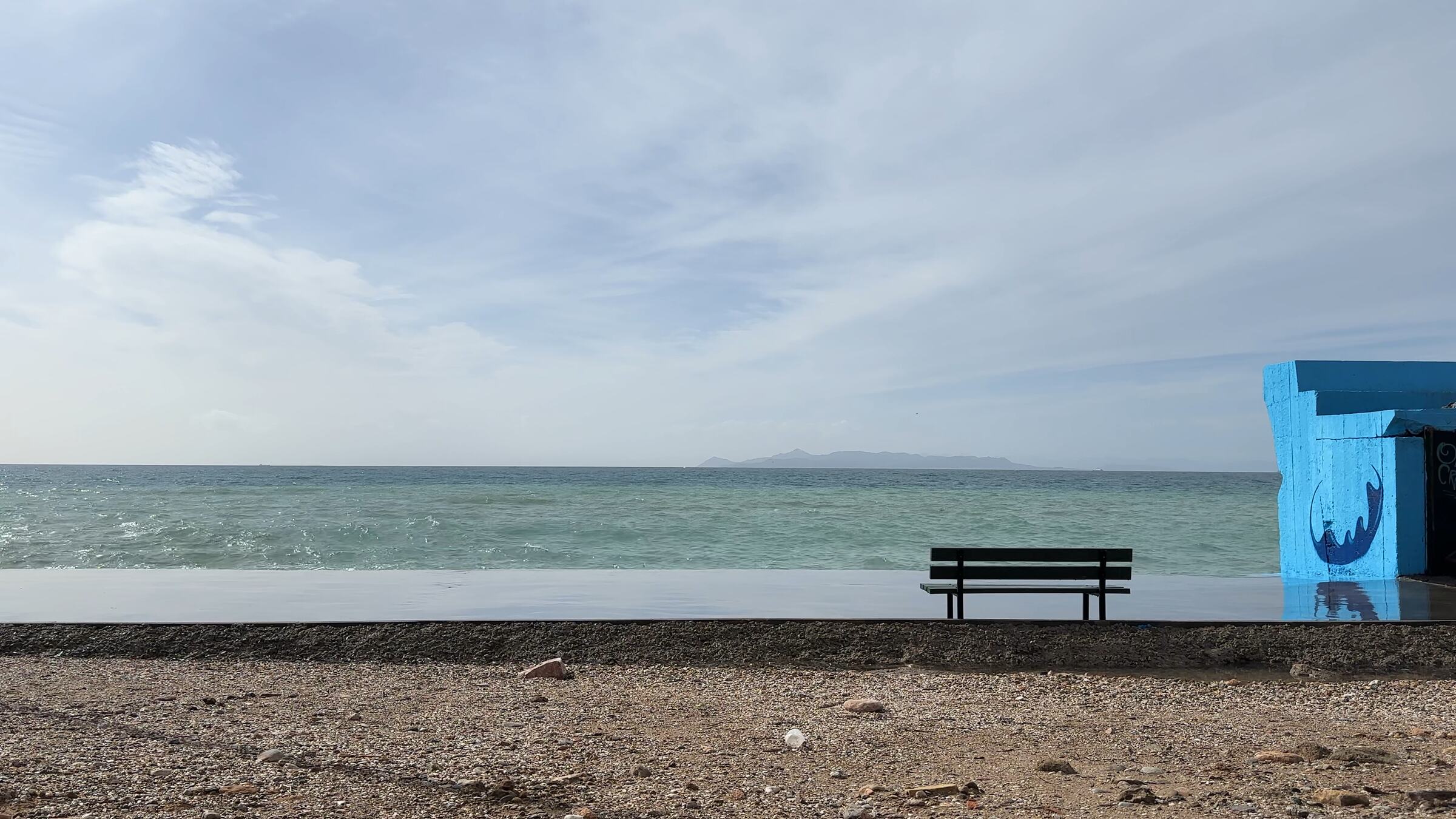 An empty bench on a wet concrete platform facing the sea at the Saronic Gulf, Athens. A faint mountain ridge on the horizon.